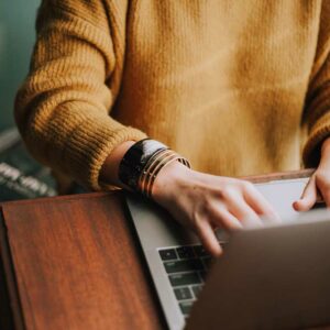 person writing on a computer in a mustard colored sweater on wood table
