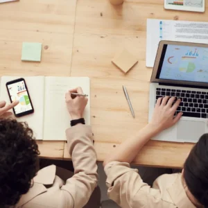 Two people working at a table on a smart phone and a laptop. The laptop screen shows graphs